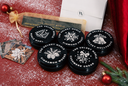 Six black circular cosmetic tins with white printed holiday tree and ornament designs are arranged on a red surface dusted with white faux snow. A white envelope, a small gold-tied pouch, and decorative cards are also visible.